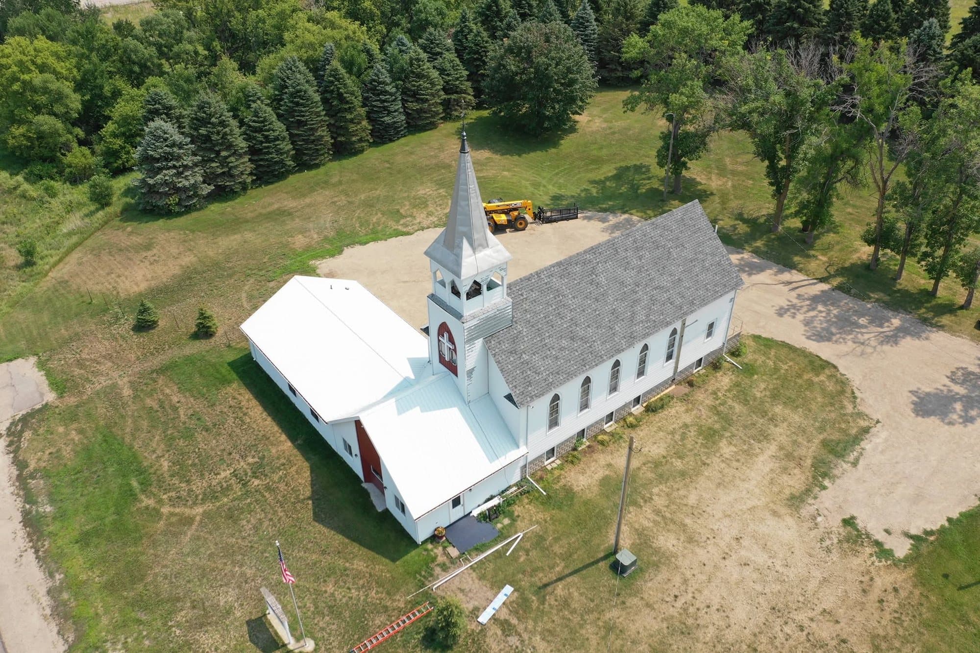 White country church with a steeple and a new gray shingle roof, surrounded by pines